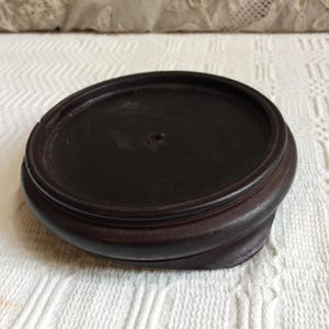 May include: A dark brown, wooden display stand with a circular top and a rounded, tiered base. The stand has a smooth, polished finish and a small hole in the center of the top surface.
