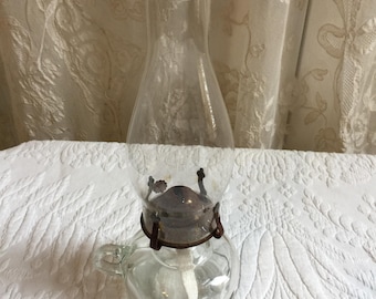 May include: A clear glass oil lamp with a metal wick holder and a glass chimney. The lamp has a handle on the side and is sitting on a white surface.