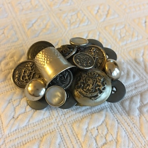 May include: A silver thimble surrounded by a cluster of vintage buttons, including a large button with a coat of arms design.
