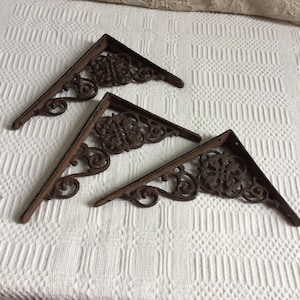 Puede incluir: Cuatro soportes decorativos de estantería de hierro fundido con un acabado marrón rústico. Cada soporte presenta un diseño floral ornamentado y volutas. Los soportes están angulados y diseñados para soportar estantes, añadiendo un toque vintage.