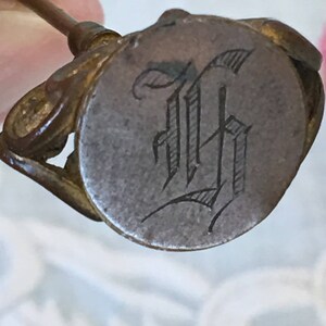 May include: A vintage brass seal with a round, silver-toned face engraved with the letter "H" in a cursive script.