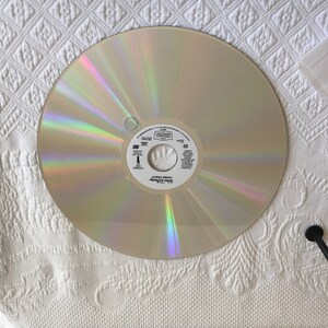 May include: A silver-colored laser disc with a rainbow iridescent sheen. The disc has a white label with the text "Disney" and "Home Video" printed on it. The text "The Little Mermaid" is printed in a larger font.