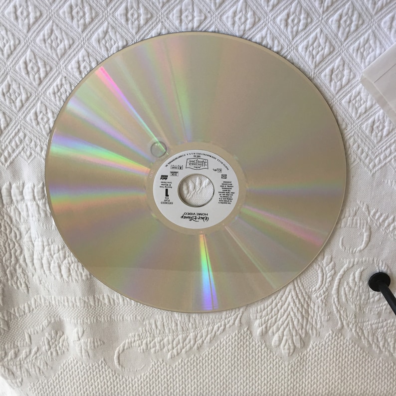 May include: A silver-colored laser disc with a rainbow iridescent sheen. The disc has a white label with the text "Disney" and "Home Video" printed on it. The text "The Little Mermaid" is printed in a larger font.