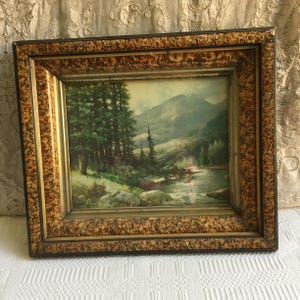 Può includere: Un dipinto di paesaggio incorniciato che rappresenta una scena di montagna con un fiume che scorre attraverso una foresta. La cornice è in legno marrone scuro con accenti dorati.