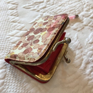 Op de afbeelding: Vintage portemonnee met een crèmekleurige achtergrond en een patroon van bruine en roze vlekken. De portemonnee heeft een rode binnenkant en een goudkleurig metalen frame met een sluiting. De portemonnee is open en onthult de binnenkant.