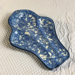 Op de afbeelding: Een keramieken lepelhouder met een onregelmatige, langwerpige vorm en een blauw-witte gespikkelde sponsafwerking. Het object heeft een klein rond gat aan de bovenkant om op te hangen en is voorzien van een gladde, glanzende glazuurlaag.