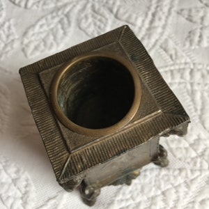 May include: A square, brass, decorative container with a round opening at the top. The container has a textured surface and appears to be antique.