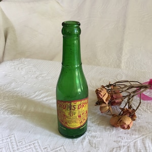 Könnte beinhalten: Eine grüne Glasflasche mit einem gelben und roten Etikett, auf dem "Rums Dry Ginger Ale" steht. Die Flasche steht auf einer weißen Oberfläche mit getrockneten Rosen.