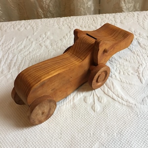Könnte beinhalten: Ein hölzernes Spielzeugauto mit einem Schlitz oben für Münzen. Das Auto ist aus hellem braunem Holz und hat vier Räder. Das Auto ist in gutem Zustand und hat einen Vintage-Look.