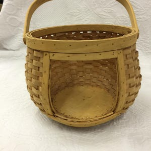 May include: A light brown, woven basket with a rounded base and a handle. The basket is made of thin strips of wood, with a sturdy construction. The handle is arched and attached to the rim. The basket has an open design, suitable for carrying items.