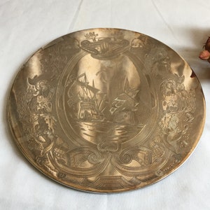 May include: A silver-toned metal plate with an engraved design featuring two ships sailing on the water. The plate is surrounded by a decorative border with a crest at the top.