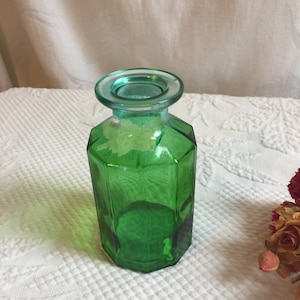 May include: A green glass bottle with a wide mouth and a faceted design. The bottle is empty and has a light blue tint.