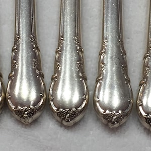 Puede incluir: Primer plano de varias cucharas plateadas con diseños florales ornamentados en los mangos. Las cucharas tienen una superficie reflectante, y los detalles de los patrones florales son visibles. Las cucharas están dispuestas en fila.