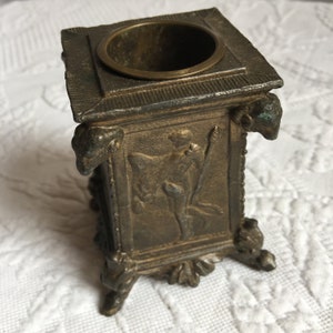 May include: A small, square, bronze inkwell with a decorative design featuring a figure with wings and a staff. The inkwell has four legs and a round opening at the top.