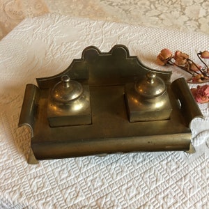 May include: A brass inkwell with two ink pots and a decorative back. The inkwell has a rectangular base and a raised back with a scalloped edge.