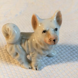 Puede incluir: Pequeña figura de perro de cerámica. El perro es blanco con orejas marrones y una nariz rosada. Tiene ojos negros y una cola rizada. La figura es detallada y parece vintage.