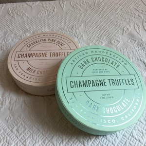 May include: Two round boxes of champagne truffles. One is light pink with "Sparkling Pink Rose" and "Milk Chocolate" text. The other is mint green with "Dark Chocolate" text. Both boxes say "Champagne Truffles" and "Artisan Handcrafted".