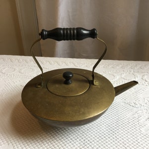 Może przedstawiać: Mosiężny czajnik z drewnianym uchwytem i pokrywką. Czajnik ma okrągły korpus i dzióbek po boku.