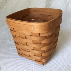 May include: A square, light brown woven basket with a slightly flared shape. The basket is constructed from thin strips of wood, with a sturdy rim and metal fasteners. The weave pattern is visible, creating a textured surface.