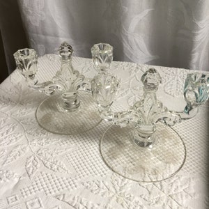 Puede incluir: Dos candelabros de cristal transparente con tres brazos cada uno. Los candelabros tienen un diseño decorativo y están sobre una superficie blanca.