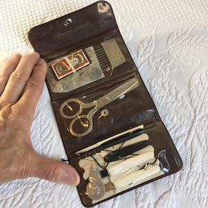 Op de afbeelding: Een bruine vinyl naaigarnituur etui met een zilverkleurige metalen schaar, een klein pakje naalden met het label 'Eagle' en diverse witte en zwarte garenklosjes. De set is voorzien van doorzichtige plastic vakjes voor de naaibenodigdheden.