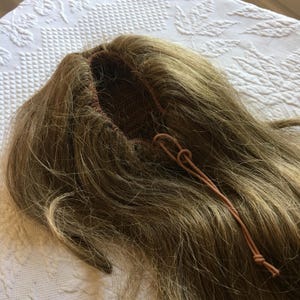 Puede incluir: Peluca marrón claro con cabello largo y liso. La peluca tiene una gorra de malla marrón y un cordón marrón. El cabello es una mezcla de tonos marrones claros y oscuros, lo que le da un aspecto natural. La peluca está sobre una superficie blanca.