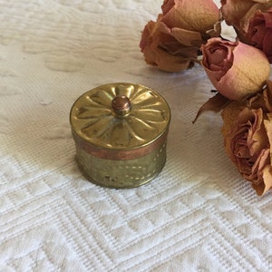 May include: A small, round, brass trinket box with a copper band around the middle. The lid has a floral design and a small knob on top. The box is sitting on a white, textured surface.