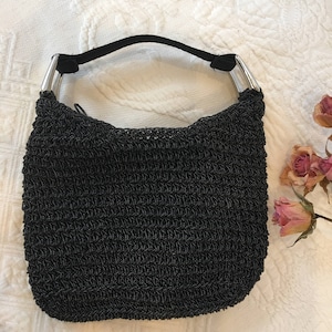 Könnte beinhalten: Eine schwarze, gehäkelte Handtasche mit silbernen Metallbeschlägen. Die Tasche hat eine gewebte Textur und eine lässige Form.