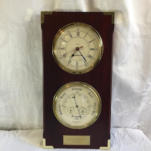 May include: A rectangular dark wood wall plaque featuring a gold-rimmed quartz clock with Roman numerals above a gold-rimmed barometer with weather indicators. A brass plate at the bottom reads EMOGENE & EATON 25 YEARS. Brass corner accents are visible.