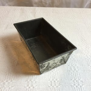 May include: A rectangular, vintage metal loaf pan with a dark, aged finish. The pan has a slightly distressed appearance, with visible wear and tear, suggesting its age and use. The interior is dark, and the exterior shows signs of oxidation.