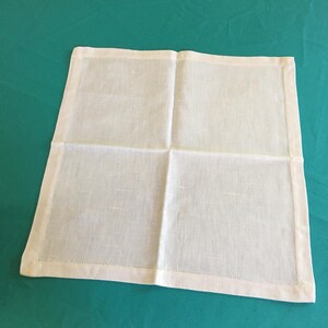 Puede incluir: Una servilleta cuadrada de lino blanquecino con un borde cosido. La servilleta está doblada en cuatro secciones iguales, con costuras visibles. La tela tiene una textura sutil y un color suave y neutro, adecuada para diversas mesas.