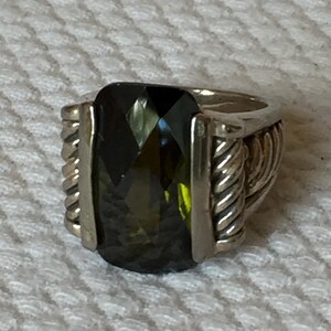 May include: Silver ring featuring a large, faceted, rectangular green gemstone. The ring has a thick band with rope-like detailing on the sides and smooth silver accents framing the stone. The gemstone reflects light, showcasing its multiple facets.