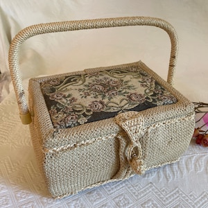 May include: A woven wicker sewing box with a floral tapestry lid and a woven handle. The box is beige and the tapestry is brown with pink and white flowers.
