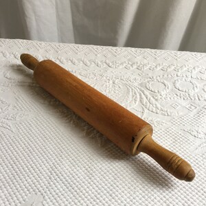 May include: A wooden rolling pin with a light brown color and a slightly darker brown handle. The rolling pin is lying on a white surface.