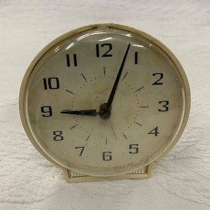 May include: A white vintage alarm clock with black numbers and hands. The clock face reads "12, 1, 2, 3, 4, 5, 6, 7, 8, 9, 10, 11" and has a small black dot at the center. The clock has a clear plastic cover over the face. The clock is made by "Columbia Gift Products LaSalle, Ill. U.S.A."