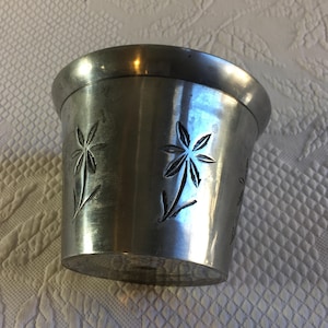 Op de afbeelding: Een zilverkleurige metalen beker met een uitlopende rand en een cilindrische body. De beker heeft geëtste bloemmotieven. De beker heeft een licht verouderd uiterlijk, wat suggereert dat hij vintage of antiek kan zijn.