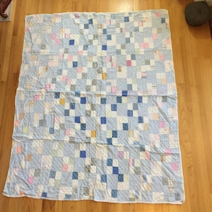 May include: A rectangular patchwork quilt featuring a grid of small fabric squares in various shades of light blue, white, pink, and yellow. The quilt displays a mix of solid colors and subtle checkered or floral patterns throughout the design.