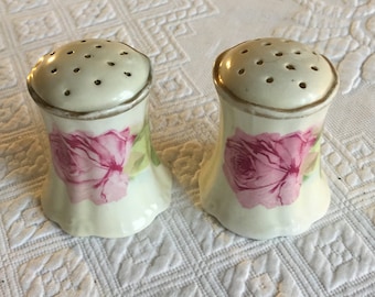 Puede incluir: Un par de saleros y pimenteros vintage con fondo blanco y diseño de rosas rosas. Los saleros tienen un borde dorado alrededor de la parte superior.