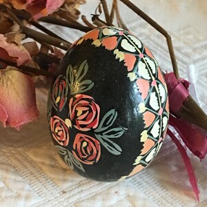 May include: A black Easter egg with hand-painted floral designs in shades of pink, red, green, and white. The egg is decorated with a geometric pattern of white and pink.