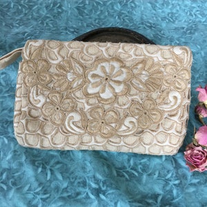 May include: A beige clutch purse with a floral embroidery design. The embroidery is in a lighter beige color and features a delicate floral pattern.
