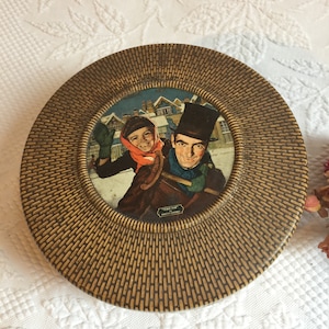 May include: A round, brown, woven tin box with a circular image of a man and a child in a snowy winter scene. The image is framed in gold and has the text "Tiny Tim" and "A Christmas Carol" printed below it.