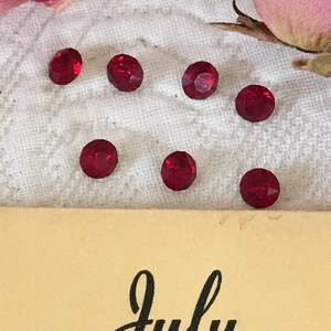 May include: Seven red glass rhinestones on a white fabric background. The rhinestones are round and have a faceted design. The background is a soft, white fabric with a subtle texture.