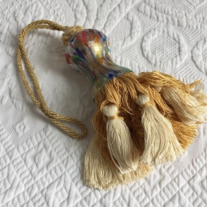 Op de afbeelding: Decoratieve kwast met een kleurrijke glazen kraal en gouden accenten. De kraal heeft rode, blauwe en gouden details. De kwast heeft een gouden koordlus en meerdere crèmekleurige kwasten.