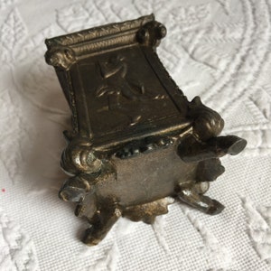 May include: A small, ornate, bronze-colored metal box with a decorative design on the front and four feet. The box is about 4 inches tall and 3 inches wide.