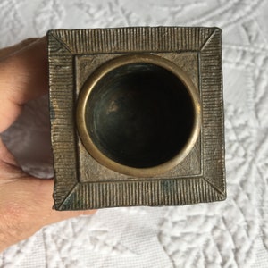 May include: A square, brass inkwell with a decorative, textured border. The inkwell has a round, open top.