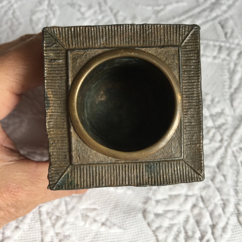 May include: A square, brass inkwell with a decorative, textured border. The inkwell has a round, open top.