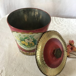 Puede incluir: Una lata de metal vintage con un diseño floral y una tapa roja con detalles dorados. La lata tiene una tapa con bisagras y un pomo.