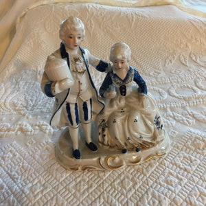 Vintage Capodimonte Colonial Pareja. Azul y blanco con estatuilla dorada. Hombre de pie sosteniendo la jaula del pájaro y mujer sentada sosteniendo el pájaro.