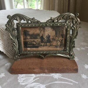 Könnte beinhalten: Ein antiker silberfarbener Bilderrahmen mit verzierten Details und einem Holzsockel. Der Rahmen enthält einen Vintage-Druck, der eine Szene mit mehreren Figuren darstellt.