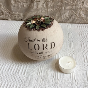 May include: Cream-colored, spherical candle holder with the text "Trust in the LORD with all your Heart" and a decorative metal flower on top. A small white tea light candle sits beside it.